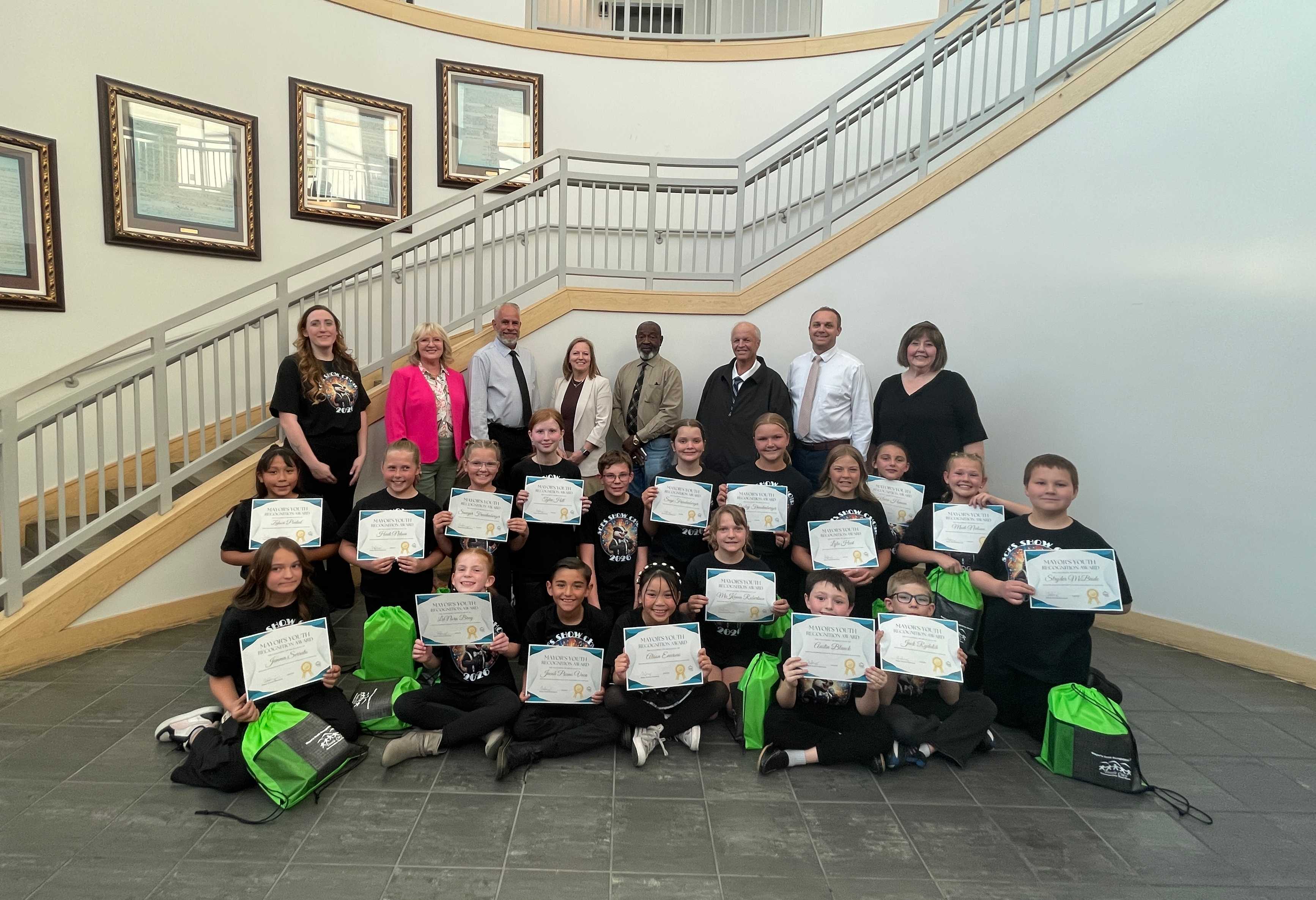 SCES Show Choir dressed in black holding a green back and sitting in front of the Mayor and City Council in the City Hall rotunda.